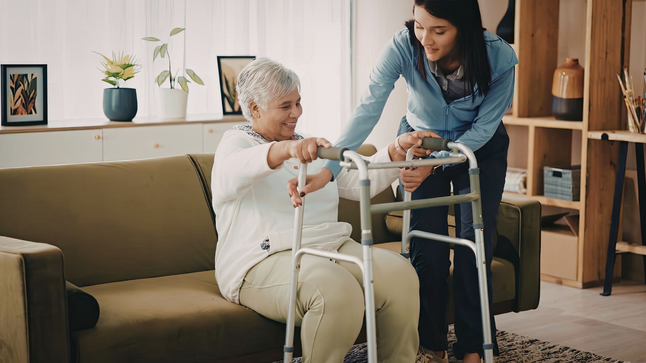 Caregiver helping elderly woman stand with a walker in her living room