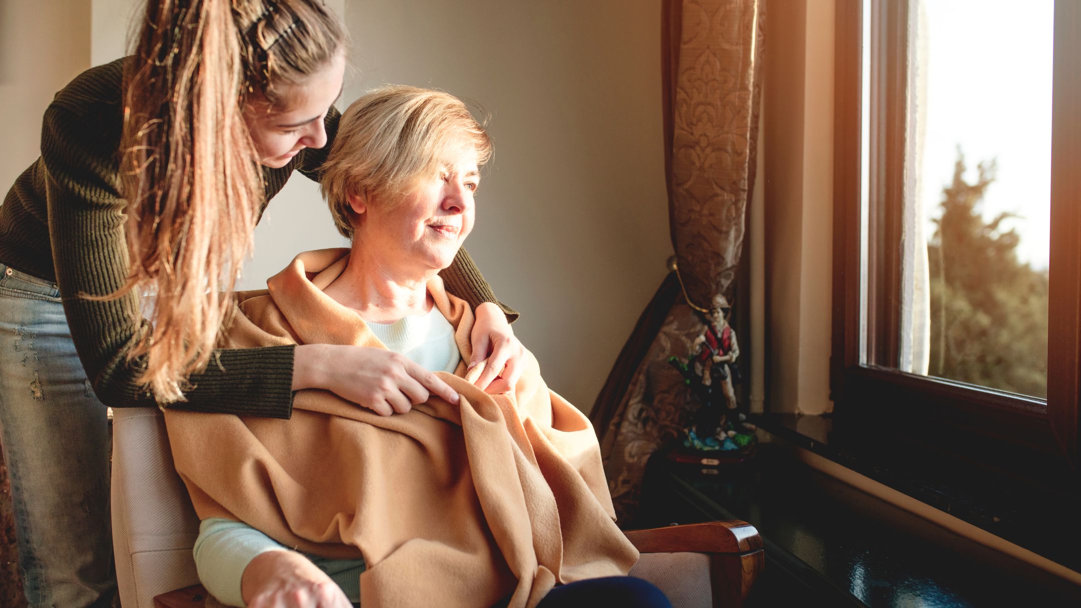 A Hearthlane caregiver wrapping a warm blanket around an elderly woman by the window